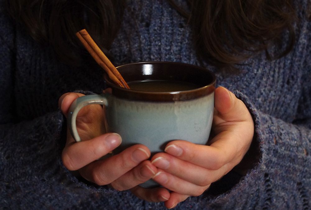 a mug of cider with a cinnamon stick, held in the hands of someone wearing a sweater
